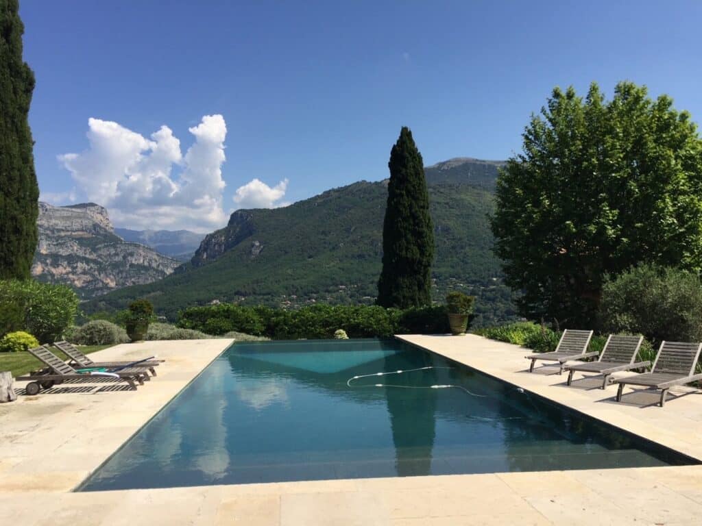 Piscine à débordement avec vue panoramique sur les montagnes verdoyantes sous un ciel bleu clair. Transats sur la terrasse ensoleillée et cyprès.