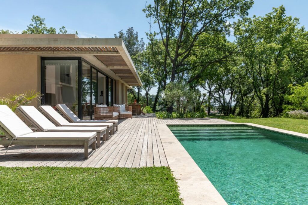 Piscine turquoise bordée d'une terrasse en bois clair, face à une maison moderne et entourée d'arbres verts luxuriants.