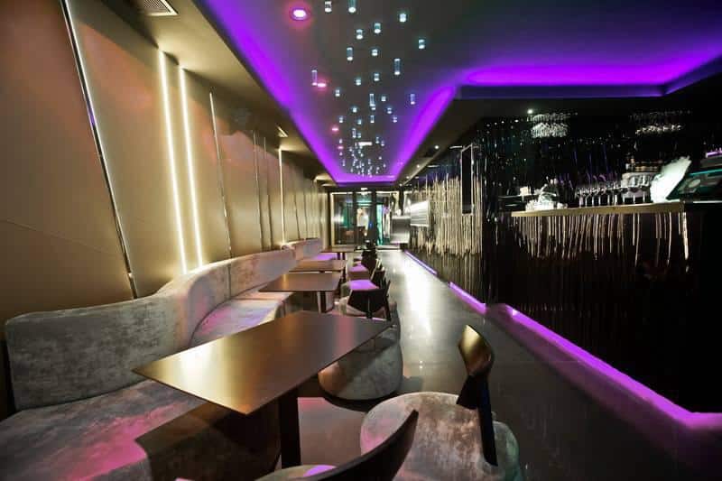 Intérieur d'un bar lounge moderne avec banquettes grises courbées, tables sombres et éclairage LED violet vif au plafond.