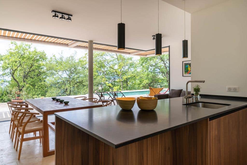 Cuisine moderne avec îlot sombre et vue panoramique sur une végétation luxuriante et une piscine extérieure.