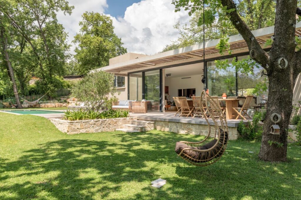 Maison moderne avec terrasse ouverte sur une pelouse verte, une balançoire en rotin et une piscine en arrière-plan, sous les arbres.
