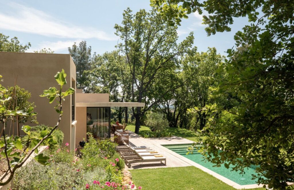 Maison moderne ocre avec piscine entourée de bois et luxuriante végétation méditerranéenne, sous un ciel bleu.