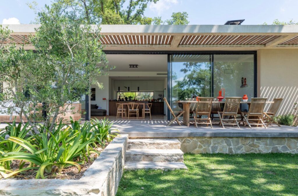 Maison moderne avec terrasse en bois, salon extérieur et grand olivier surplombant une pelouse verte et des marches en pierre.