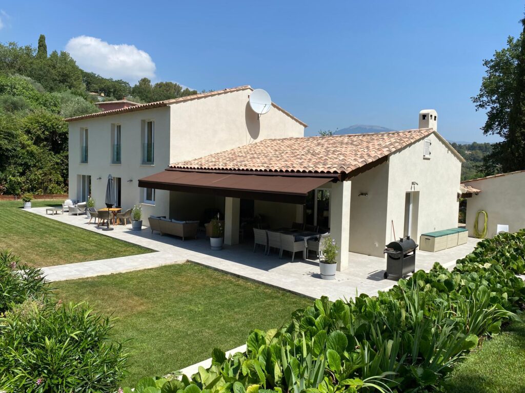 Superbe villa provençale avec toit en tuiles rouges, jardin verdoyant, terrasse couverte, mobilier extérieur et vue sur des collines boisées.