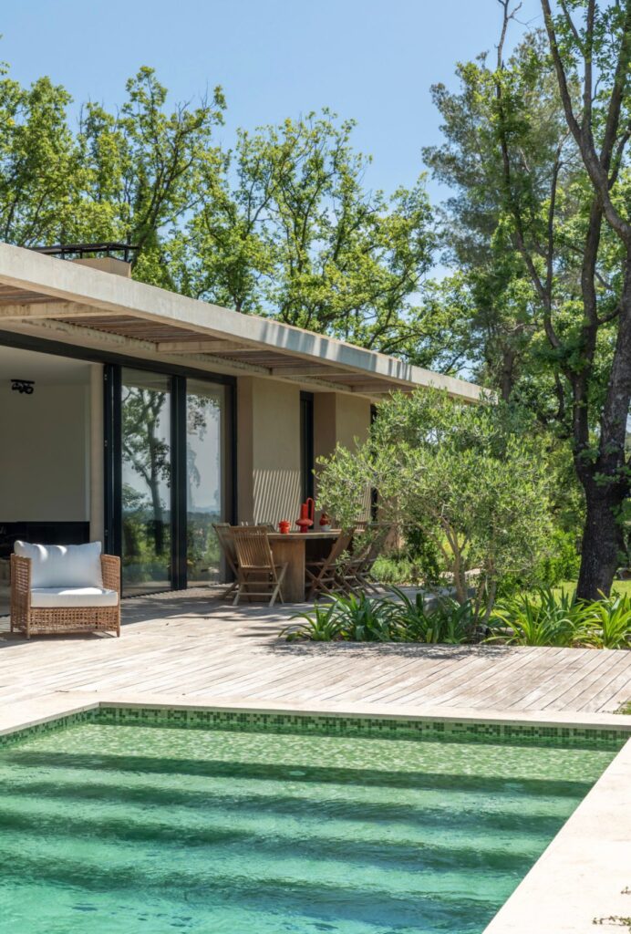 Terrasse en bois clair avec piscine aux mosaïques vertes, mobilier d'extérieur en rotin et bois sous un auvent, entourée de verdure.