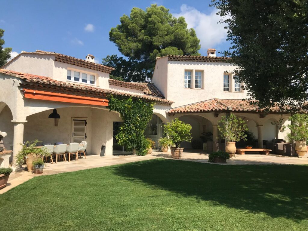 Belle villa provençale avec toits de tuiles rouges, pelouse verte bien entretenue et terrasse couverte avec mobilier d'extérieur.