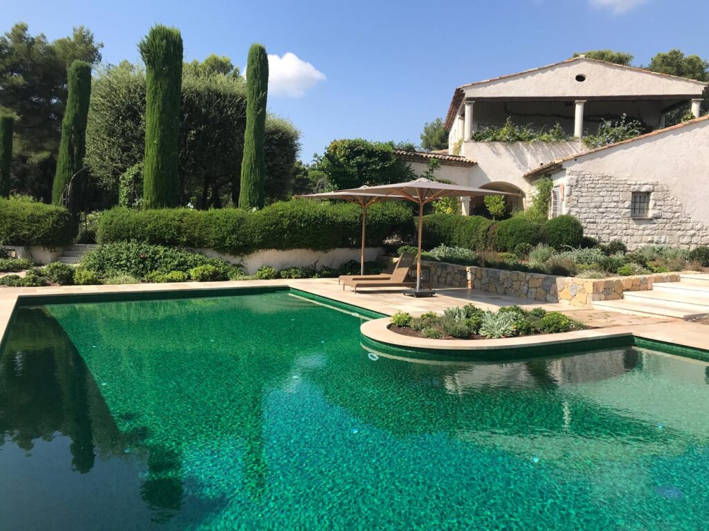 Piscine luxueuse aux carreaux vert émeraude, entourée de cyprès élancés, d'un jardin méditerranéen et d'une bastide en pierre.