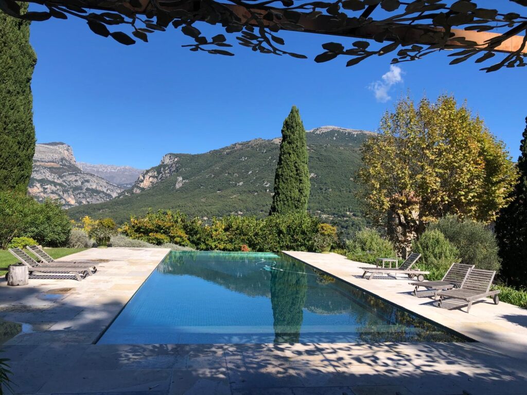 Piscine à débordement ensoleillée avec vue panoramique sur des montagnes verdoyantes et un ciel bleu clair, sous une pergola feuillue.