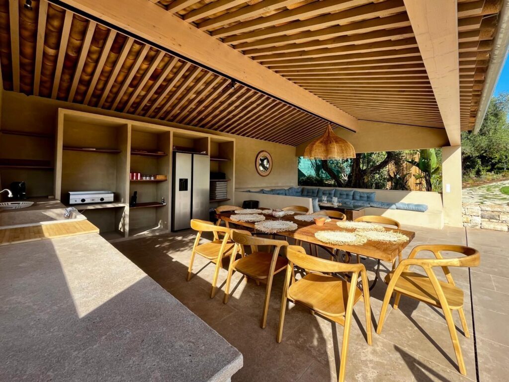 Salle à manger extérieure couverte, mobilier bois clair, cuisine intégrée rustique, lumière dorée du soleil couchant.