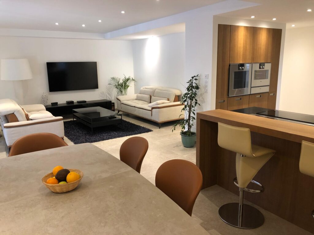 Salon moderne ouvert sur cuisine avec canapés clairs, TV murale, table à manger, et îlot bar avec tabourets en cuir.