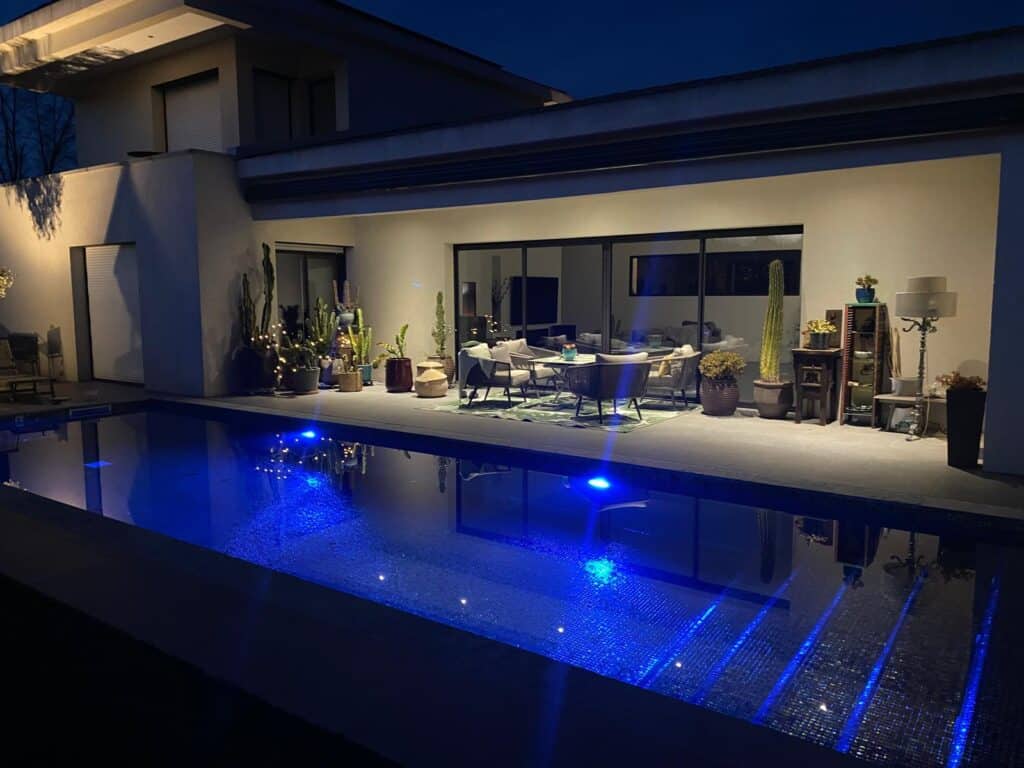 Ambiance nocturne relaxante près d'une piscine aux lumières bleues, avec terrasse moderne, salon d'extérieur et nombreux cactus éclairés.