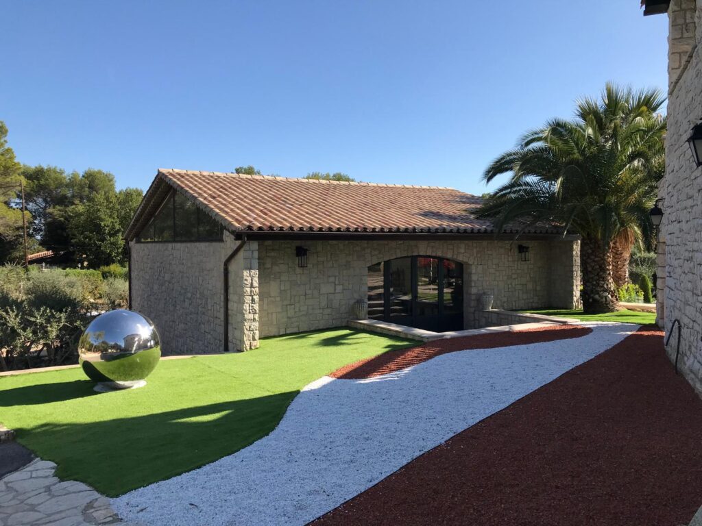 Aménagement Paysager Moderne avec Villa en Pierre Maison en pierre, pelouse synthétique, sphère miroir et allées de gravier coloré sous un ciel bleu ensoleillé.